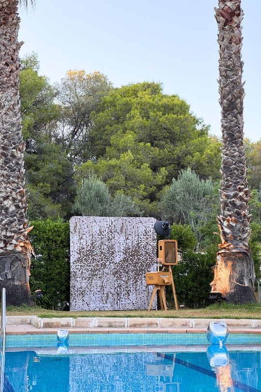 El fotomatón de madera con fondo dorado junto a la piscina de Villa Golf, entre palmeras — Sitges